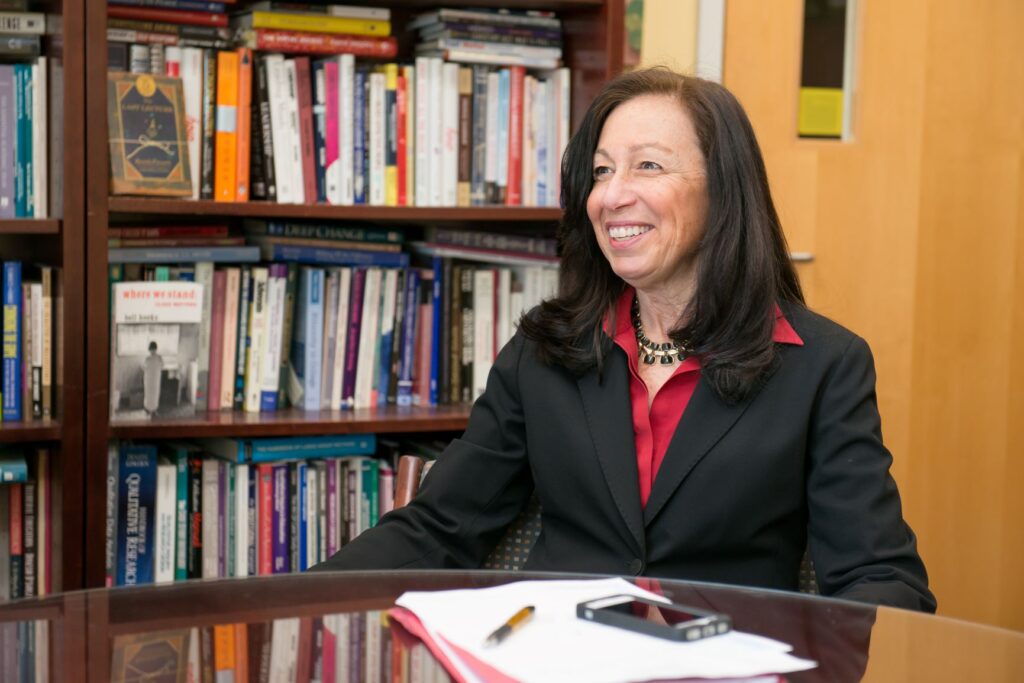 Portrait of middle-aged white woman with long, dark hair. She sits at a desk in an office, with books in the background. She wears a red collared shirt and black suit jacket.