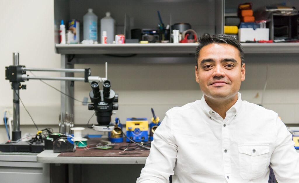 Portrait of a smiling middle-aged South Asian man wearing a white button up shirt. He sits in front of a desk with tech equipment.