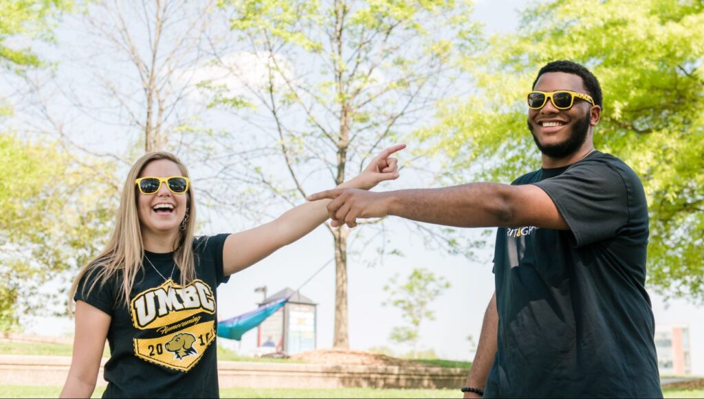 Two college students (a white woman and Black man) smile for an outdoor portrait. They point toward each other. They wear sunglasses and black and gold UMBC t-shirts.