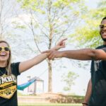 Two college students (a white woman and Black man) smile for an outdoor portrait. They point toward each other. They wear sunglasses and black and gold UMBC t-shirts.