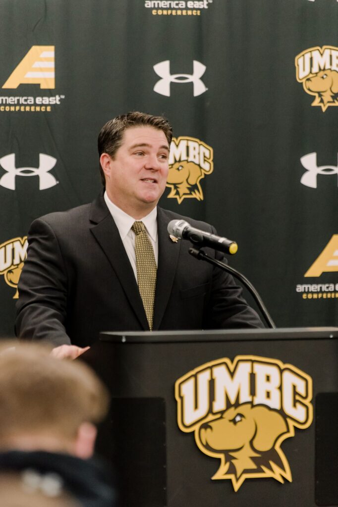 Middle-aged man with short, dark hair stands at a podium. The podium has a UMBC Athletics logo, as does a banner behind him. He wears a suit with yellow and black tie.