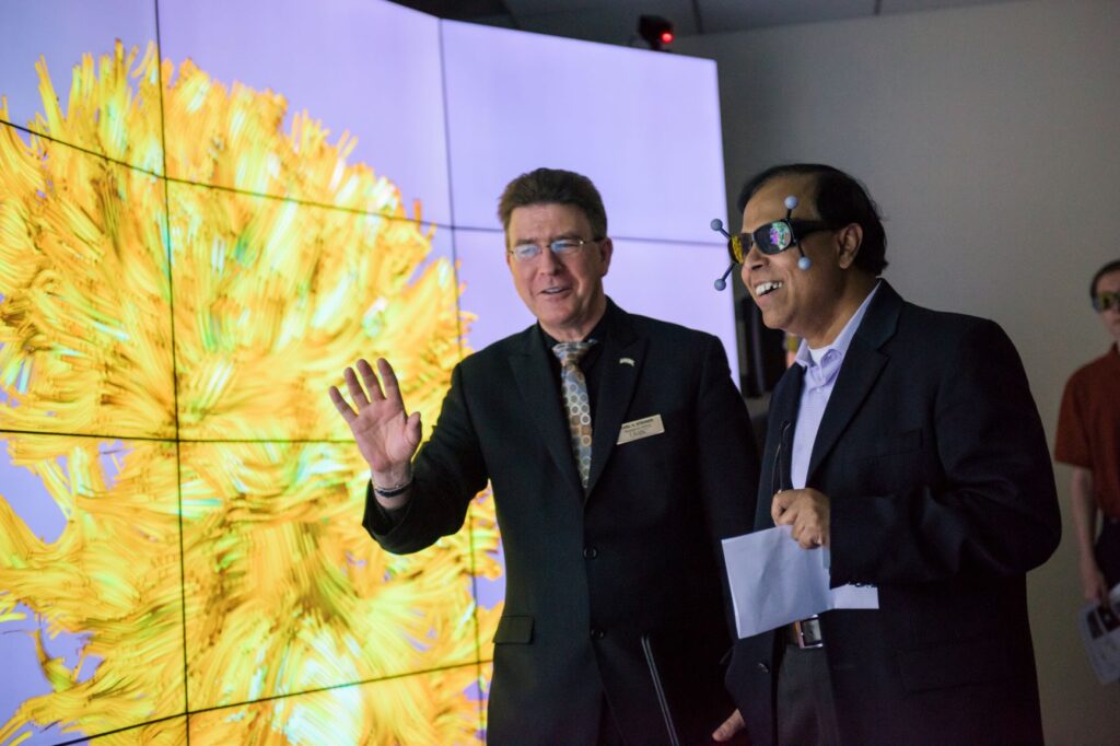 Two men in students stand in front of a brightly lit screen. One gestures while the other wears VR goggles with amazed facial expression.