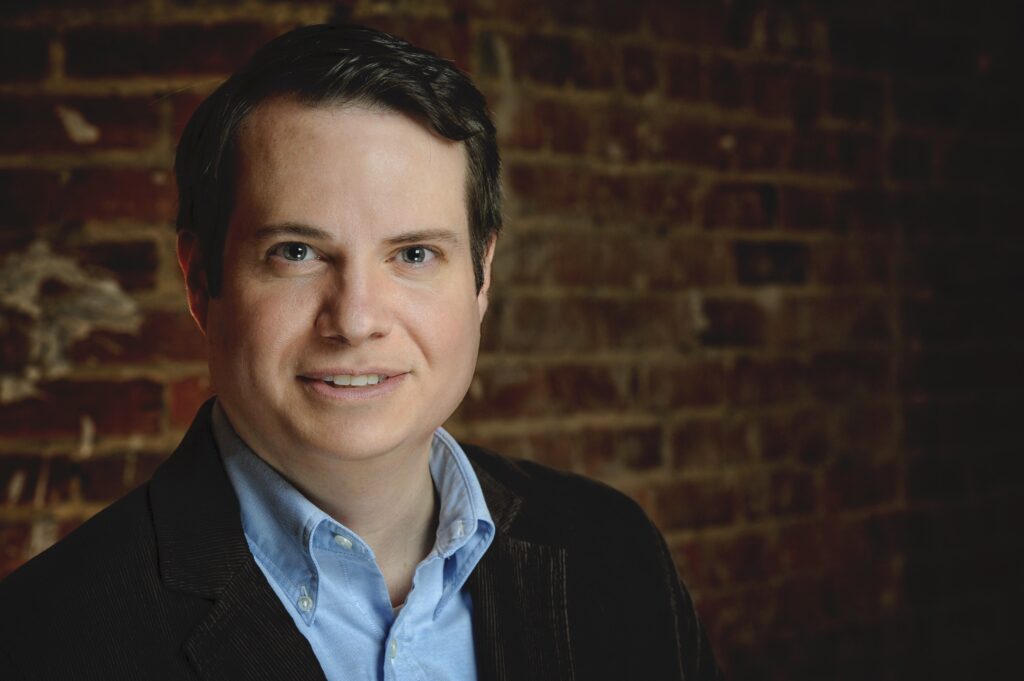 Portrait of a white man with short, dark hair in a blue shirt and suit jacket. He stands in front of a brick wall.