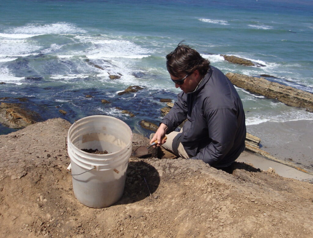 A man wearing a blue jacket sits on a cliff by the sea with shoveling  small mounds of dirt into a white bucket.