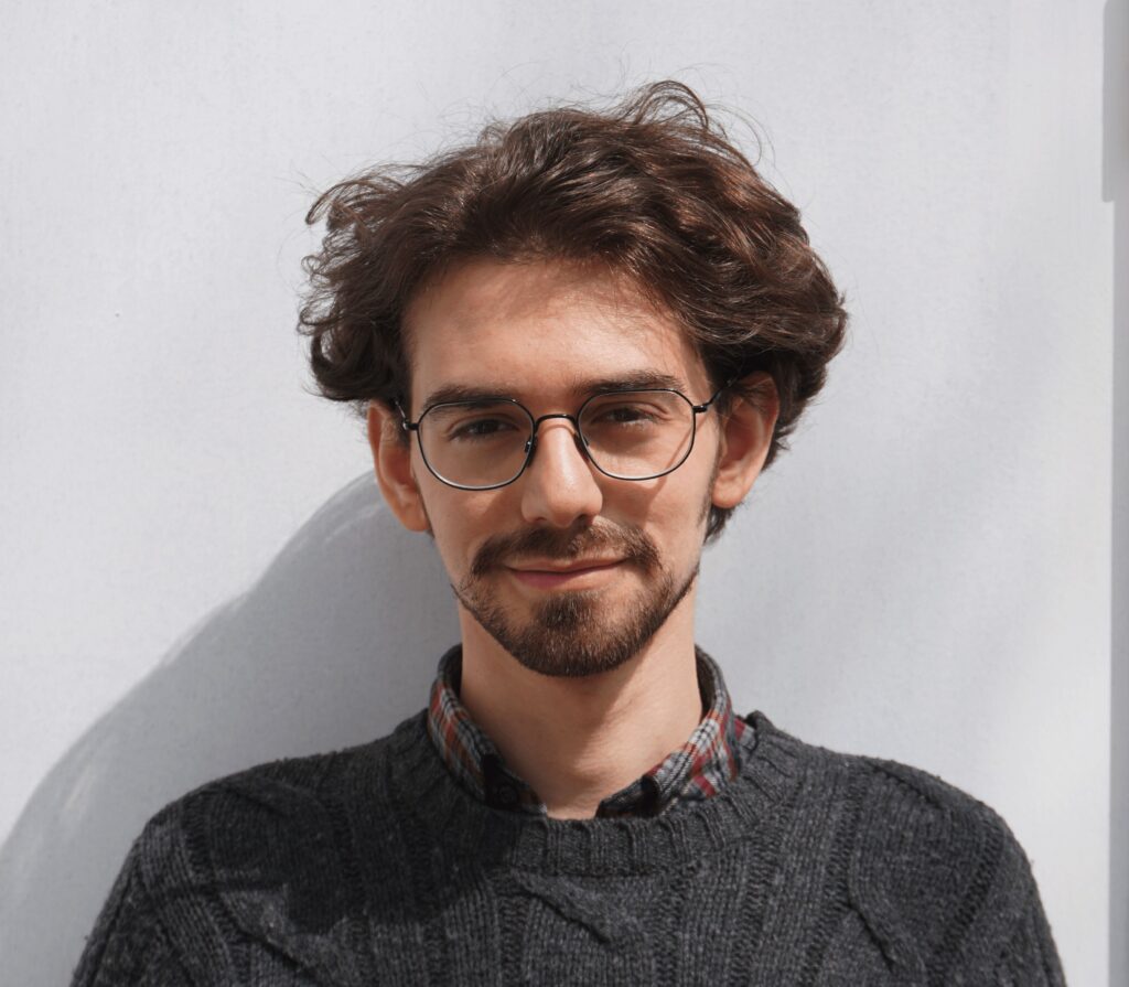 Outdoor portrait of a smiling young man with wavy brown hair, a mustache and beard. He wears a gray sweater and wire-framed glasses.