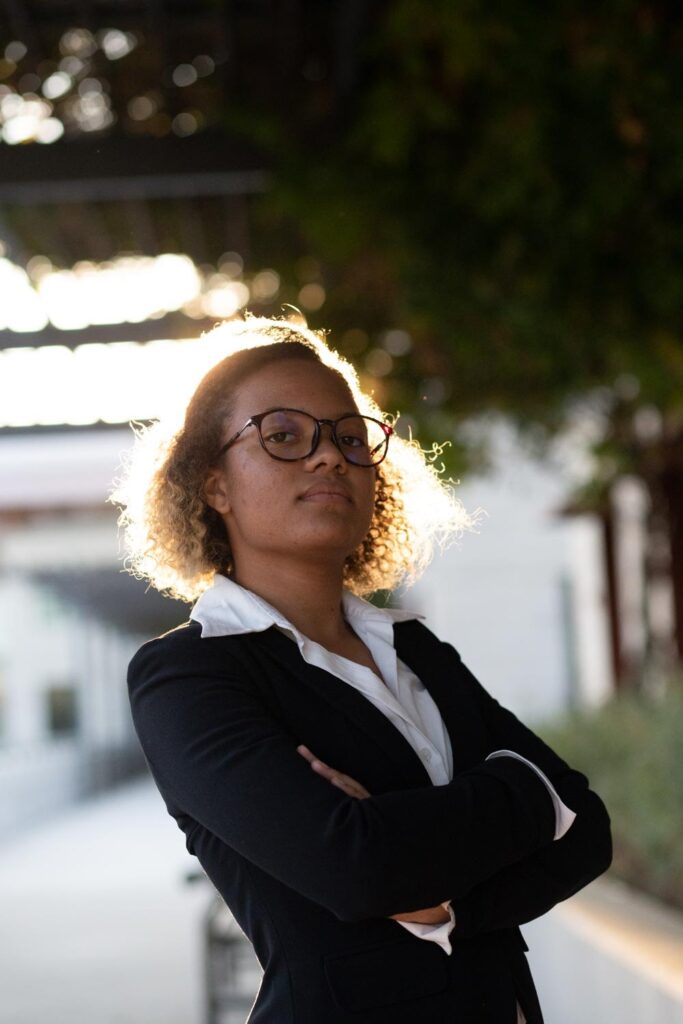 Outdoor portrait of a young black woman wearing a suit and glasses. The sunlight shines behind her.