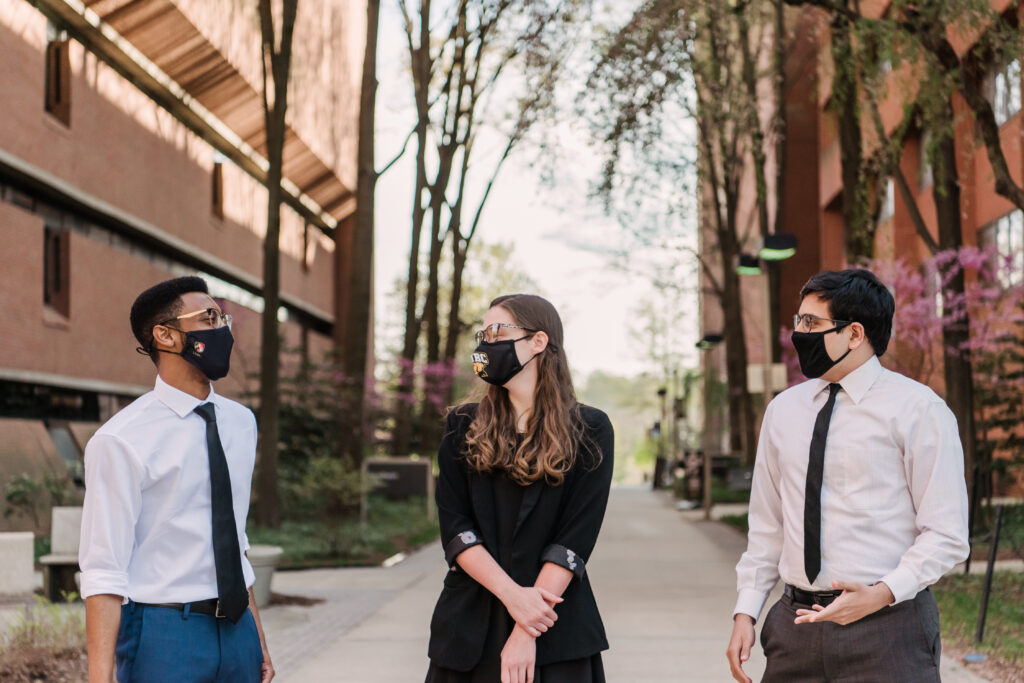 Three students (two men and one woman), all wearing masks covering their noses and mouths, stand in walkway between two brick buildings surrounded by trees.