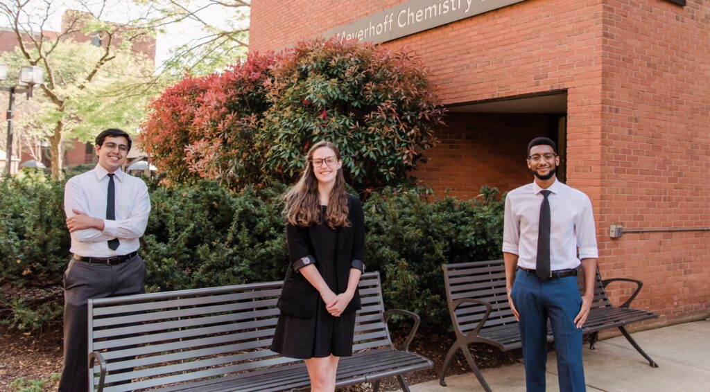 Three young adults in professional clothing stand by a park bench.