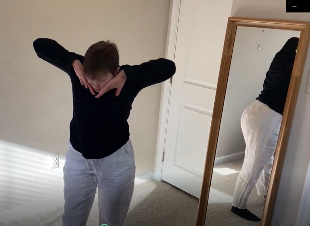 Young white woman with short brown hair touches her face with the backs of her hands in a dance. She wears a black shirt and white pants. She stands in a room with light paint and carpet, with ha mirror behind her.