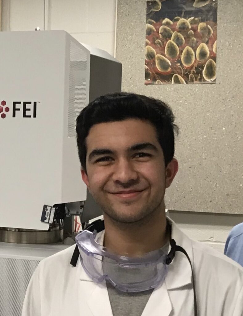 Portrait of young Middle Eastern man, smiling, wearing a lab coat and protective goggles.
