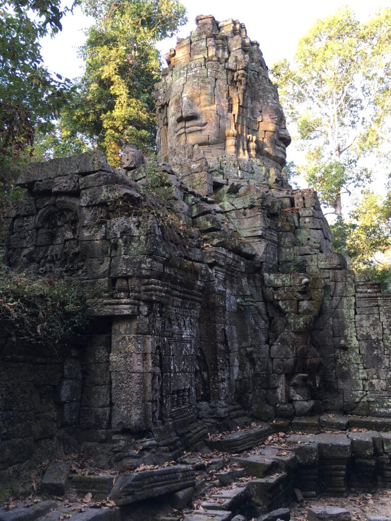 An ancient tall rock formation with a base of eroded grey rocks jutting in and out and a rock tower with two large wide faces carved into two of its sides surrounded by tall green trees.