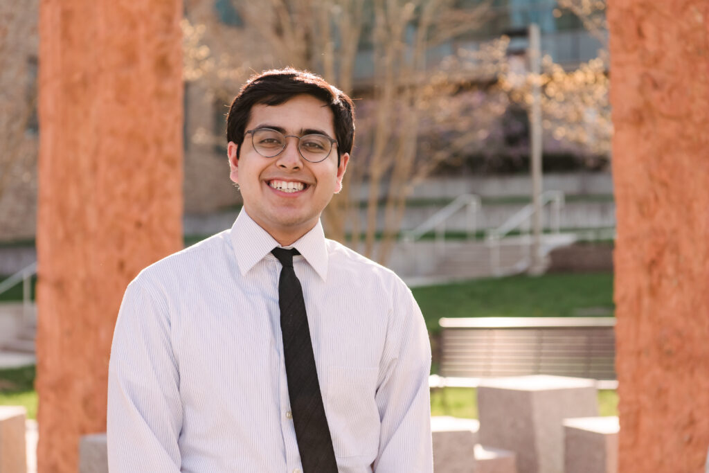A young man wearing glasses and a white shirt and dark tie smiles in front of abstract structures outdoors.