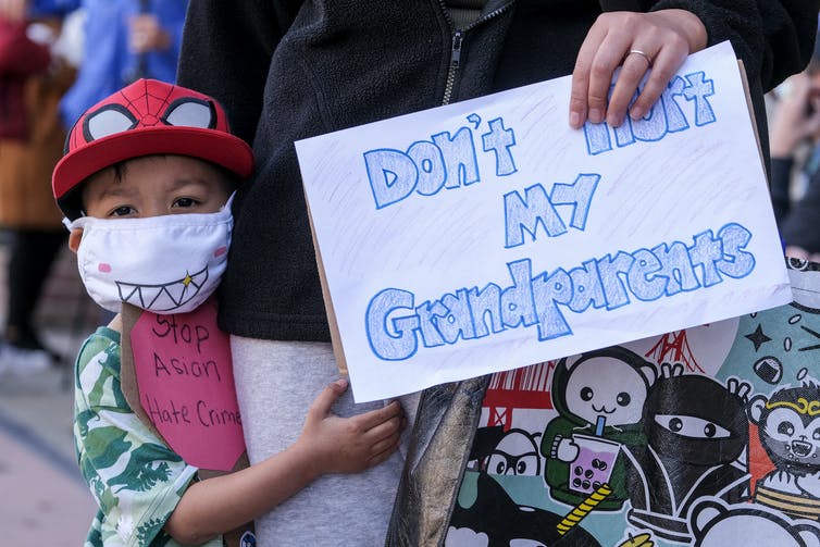 A young Asian American boy wearing a mask stands next to someone holding a sign which reads 'Don't hurt my grandparents.'