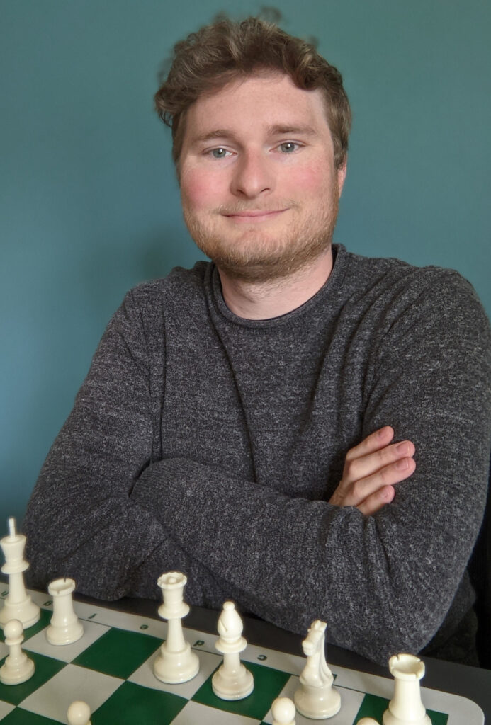 Portrait of a smiling young man in a gray sweater. He sits behind a chess board.