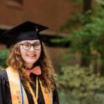Young adult with long, curly hair smiles for a portrait, wearing graduation regalia and a bow tie.