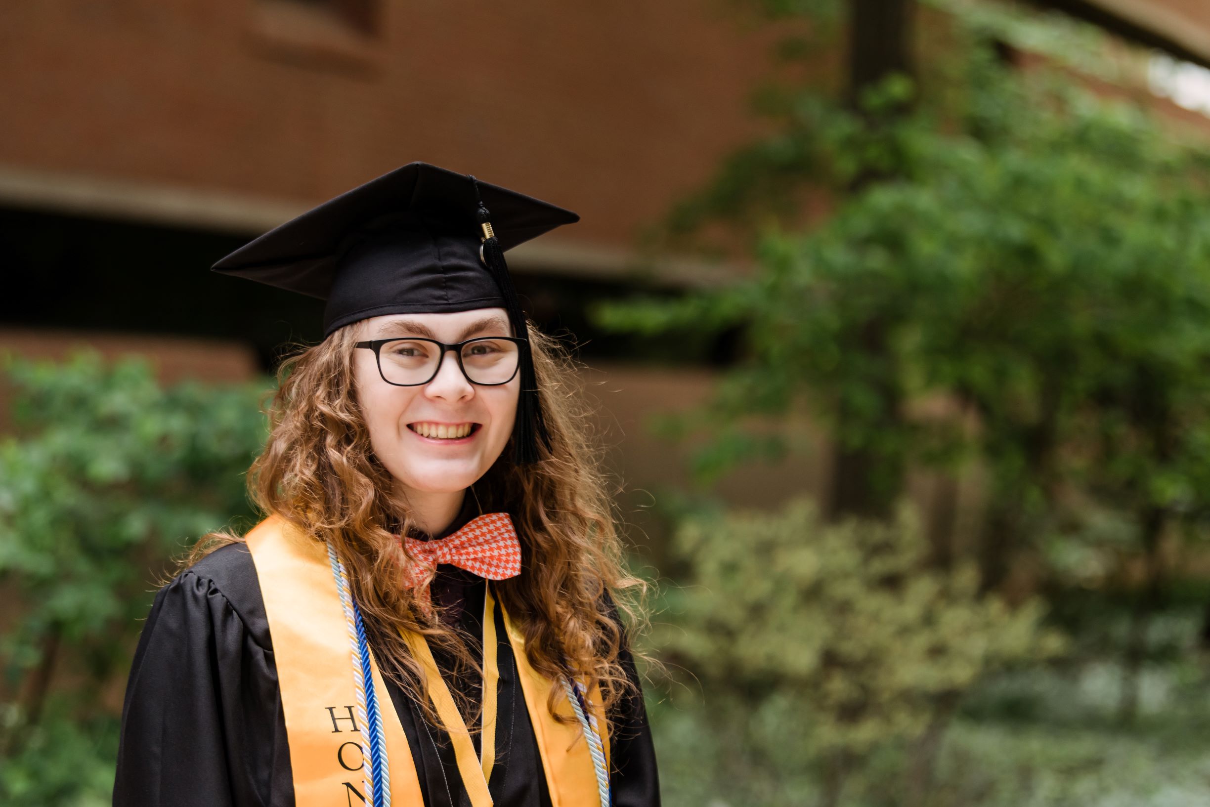 Young adult with long, curly hair smiles for a portrait, wearing graduation regalia and a bow tie.
