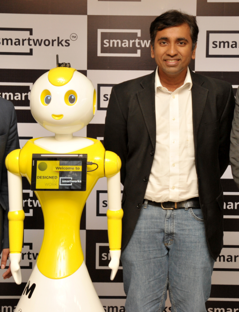 A man wearing a white shirt, dark jacket, and jeans, stands next to a yellow and white robot. There is a screen on the middle of the robot.