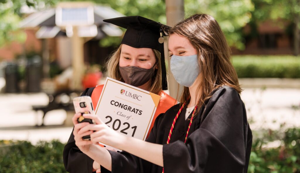 Two students, both wearing masks and graduation regalia, take a selfie. One student is holding a "Congrats Class of 2021" sign.