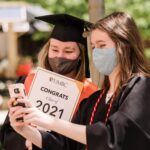Two students, both wearing masks and graduation regalia, take a selfie. One student is holding a "Congrats Class of 2021" sign.