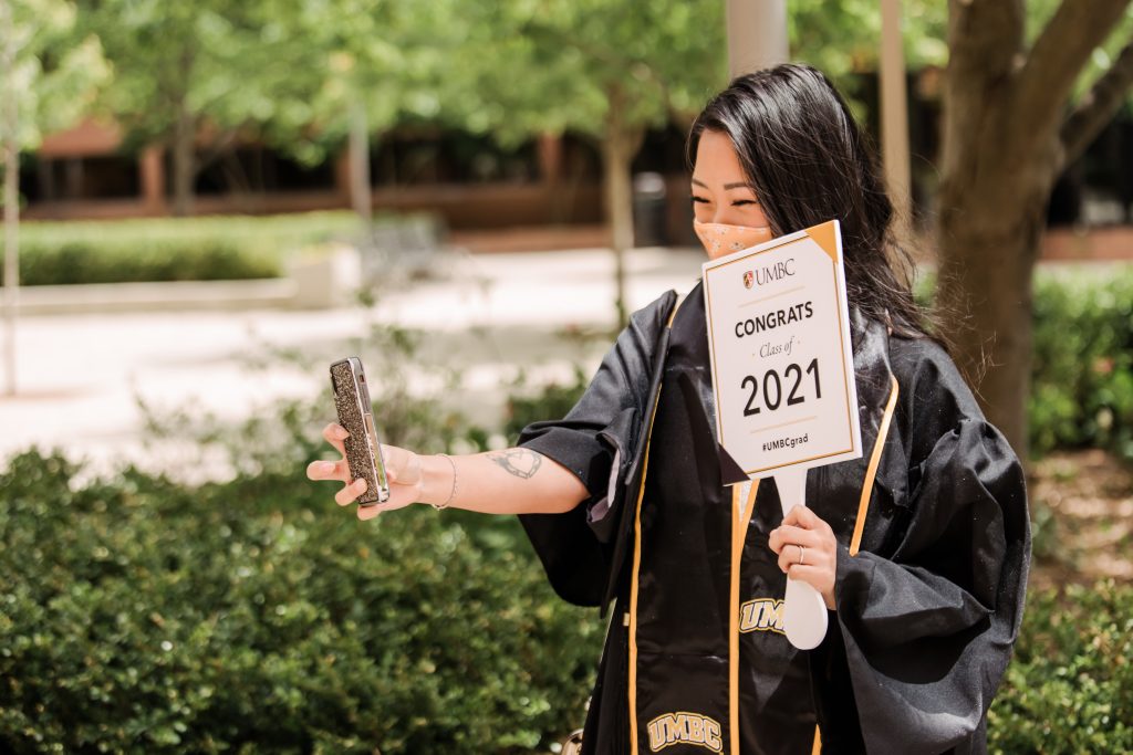 A student taking a selfie is wearing graduation regalia and a mask and is holding a sign that says "Congrats class of 2021" 