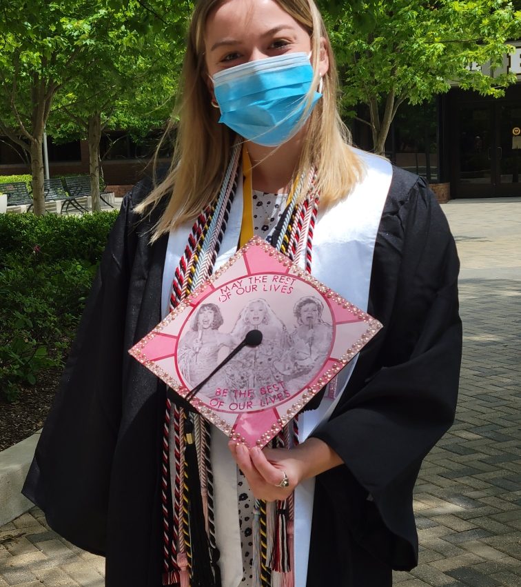 A student wears graduation regalia and a blue mask. She holds a mortar board that is decorated with a quote.