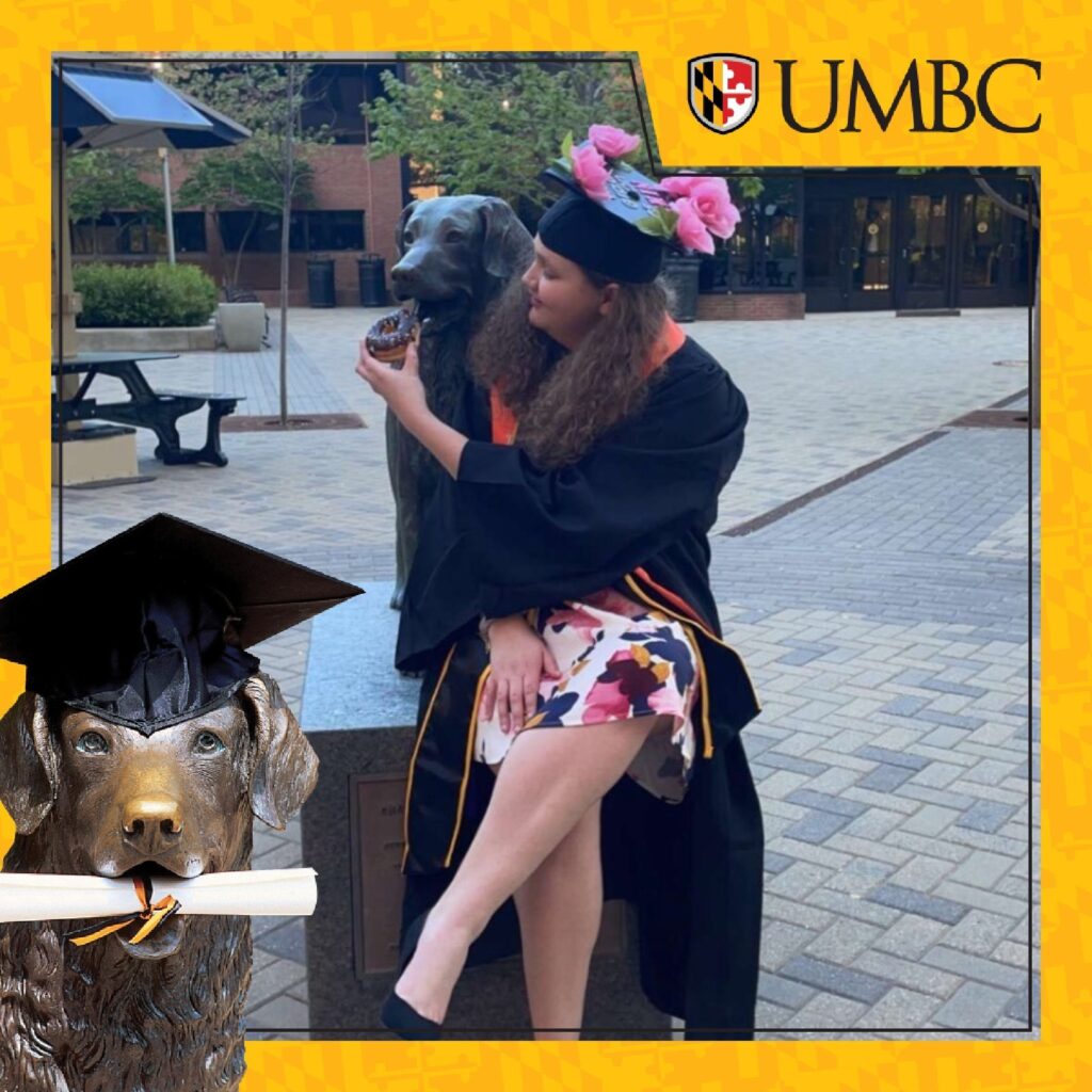 A student wearing a dress and graduating regalia, sits near the True Grit statue, and pretends to feed the statue a donut.