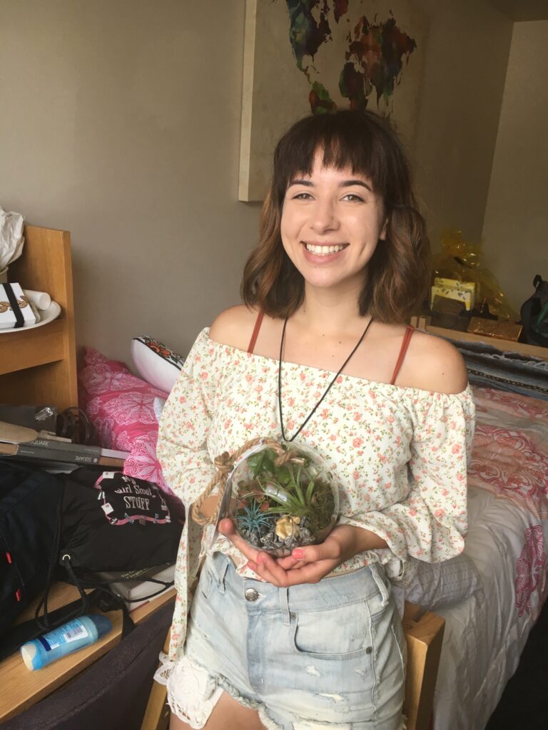 A young woman wearing an off the shoulder flow print blouse and jean shorts holds a glass bowl and smiles at the camera there is a bed and a desk in the background.