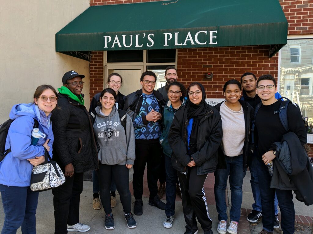 A group of eight young people with three adults wearing winter coats stand before a brick building with a green awning that says Paul's Place in white letters.