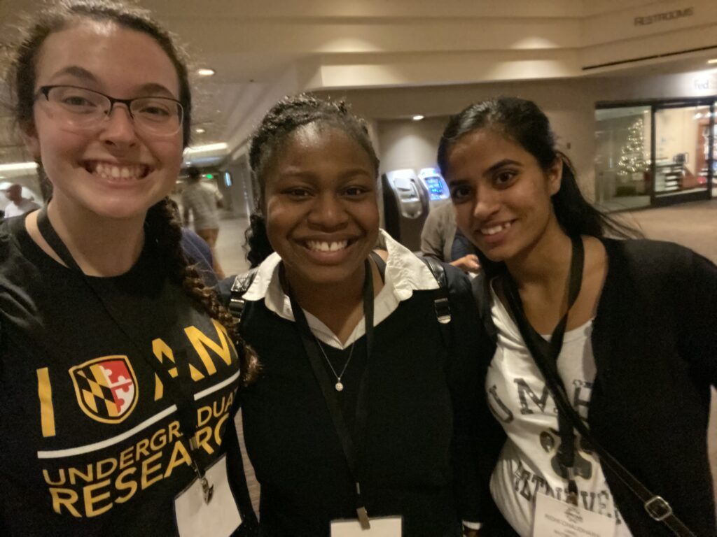 Three young women wearing lanyards around their neck stand close together and smile at the camera.