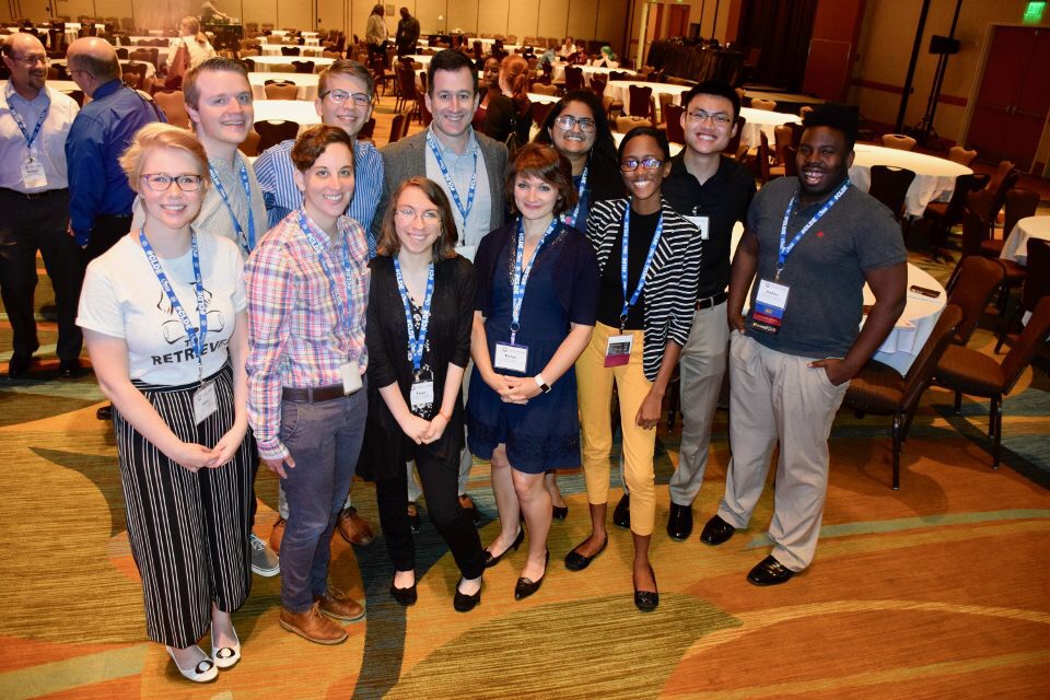 A group of 11 people in dress casual attire stands, smiling for a group photo. They wear blue conference lanyards with name tags.