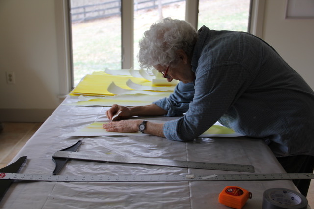 A woman with short, curly, white hair wearing a denim long sleeve shirt leans over a long table covered with white and yellow paper paper and long metal rulers.