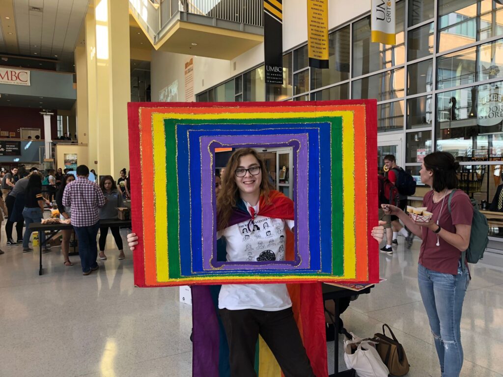 Young person stands inside a photo frame painted with rainbow colors and glitter. They wear a festive, multicolored cape.