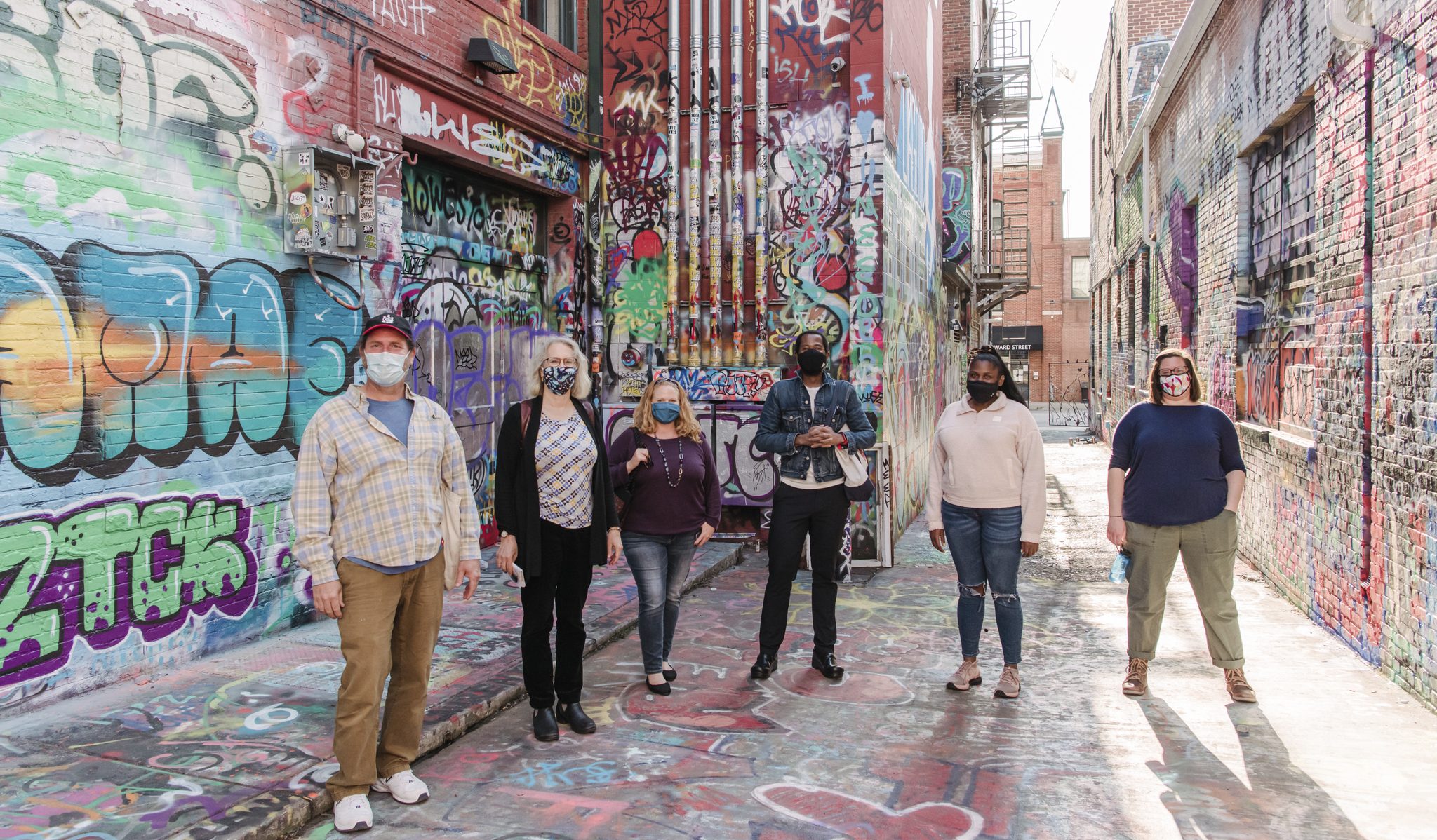 Six people, all wearing masks, standing in a road. The surrounding walls have graffiti on them.