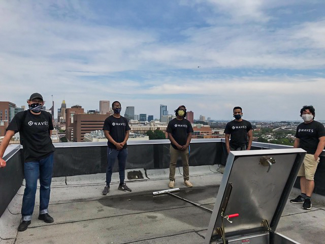 Five young men wearing black t-shirts and face masks stand on the roof of a building with a city landscape behind them.
