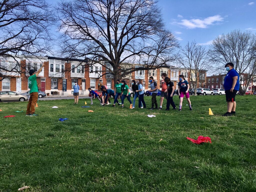 A group of children in a field of grass with row homes and bare trees in the background line up in a row facing sideways looking at a man holding his arm up.