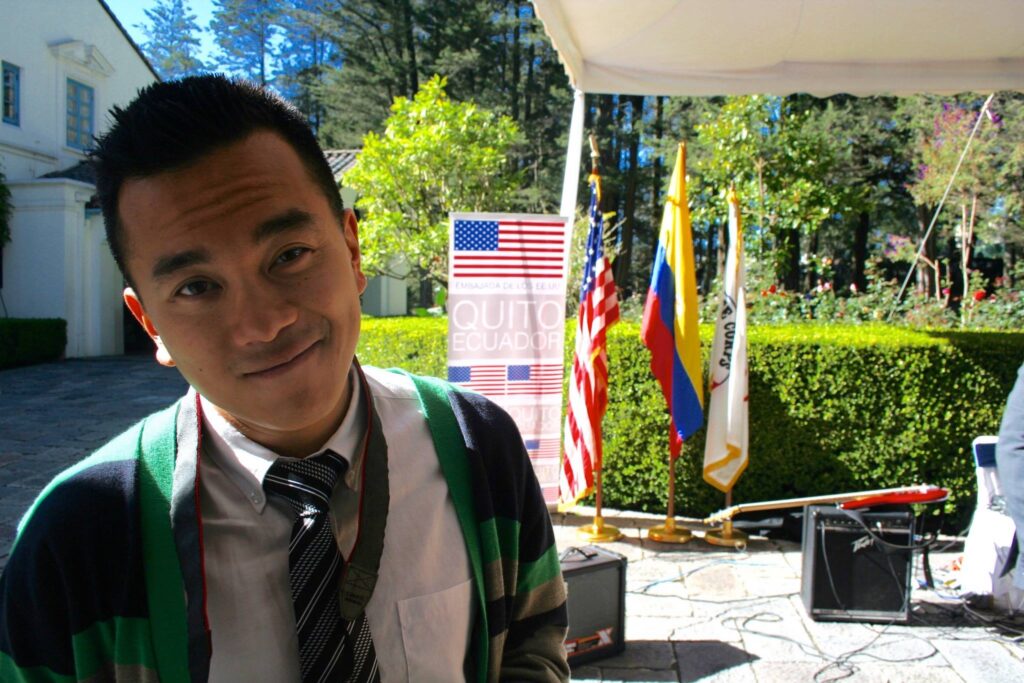 A young man with short black hair wearing a white collar dress shirt a black and white striped tie and a green, yellow, and black cardigan stands in front of three large flags of different colors.
