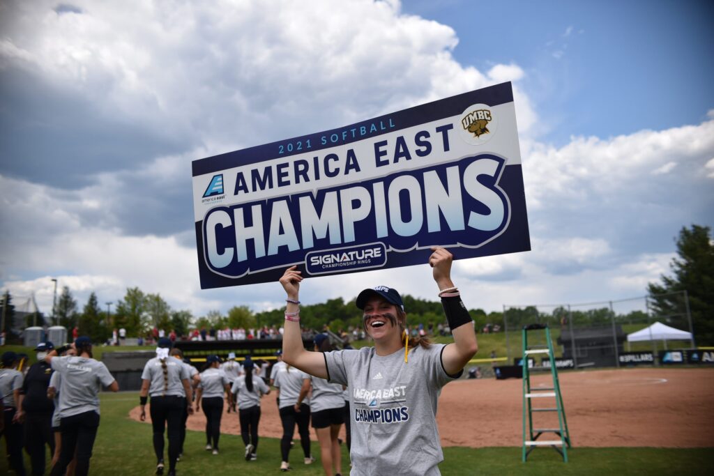 A young woman dressed in spirit-wear, smiles enthusiastically and holds up a large sign that reads "America East Champions"