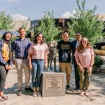 Six people stand in the sunshine, posing with a statue of a dog.