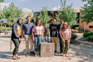 Six people stand in the sunshine, posing with a statue of a dog.