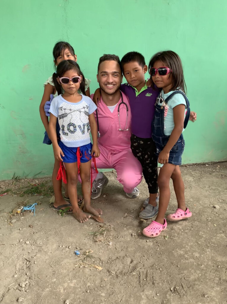 A smiling young man wearing pink scrubs and a stethoscope poses with four children, two in sunglasses.