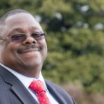 A man wearing a navy blue suit, red tie, white dress shirt, and dark rimmed glasses smiles at camera