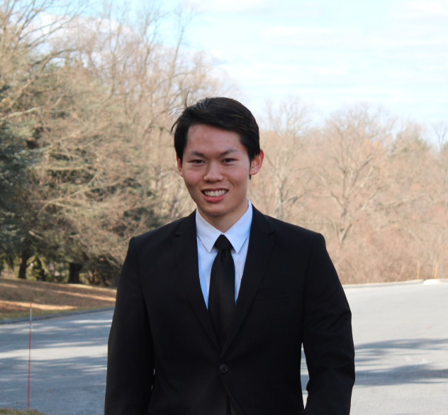A young man with short straight black hair parted to the side wearing a black suit and white dress-shirt smiles at the camera. Trees without leaves are in the background.