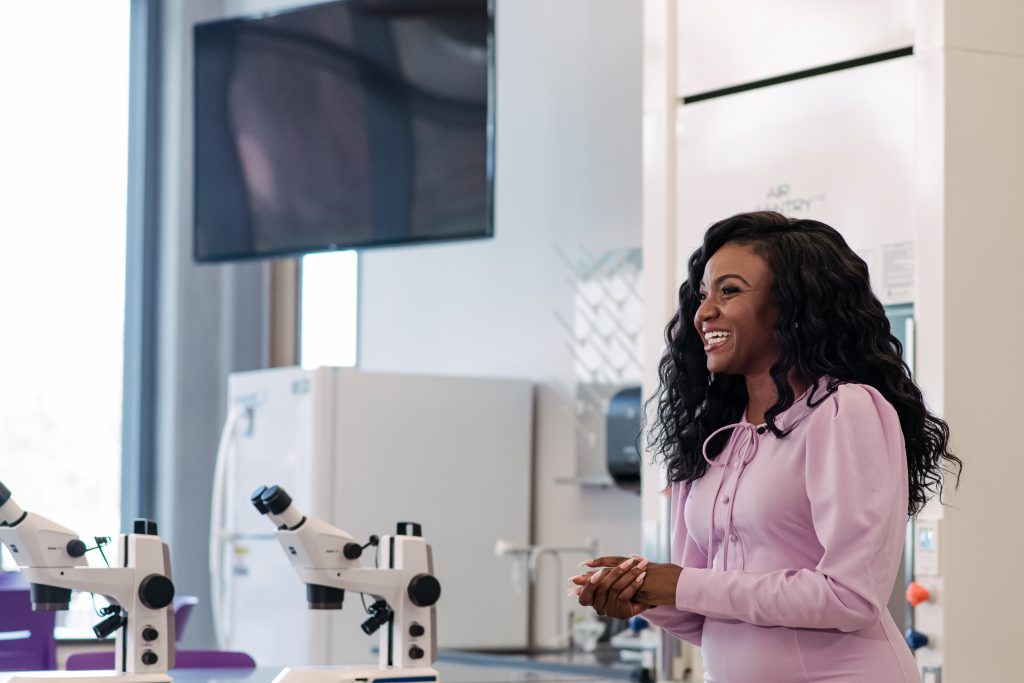 A young black woman wearing a lavender dress, smiling and standing in a lab with microscopes.
