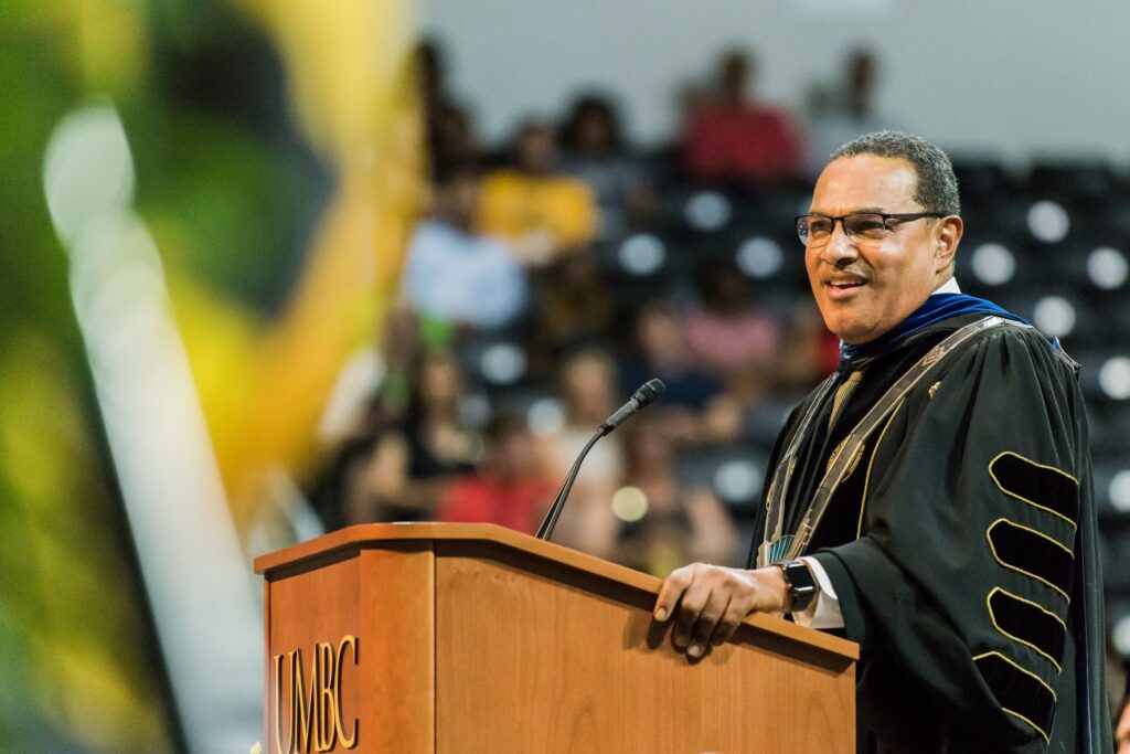 Man stands at podium in full academic regalia. Podium sign reads 