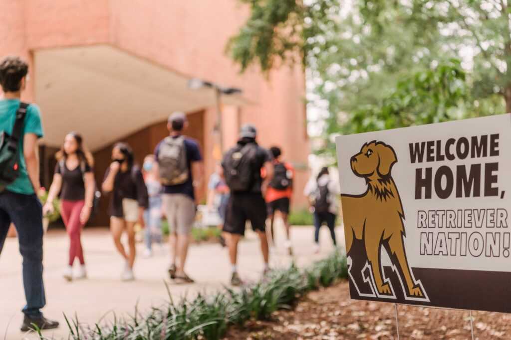 Students walk along UMBC's academic row. A sign with a graphic illustration of UMBC's mascot reads "Welcome home, Retriever nation!"