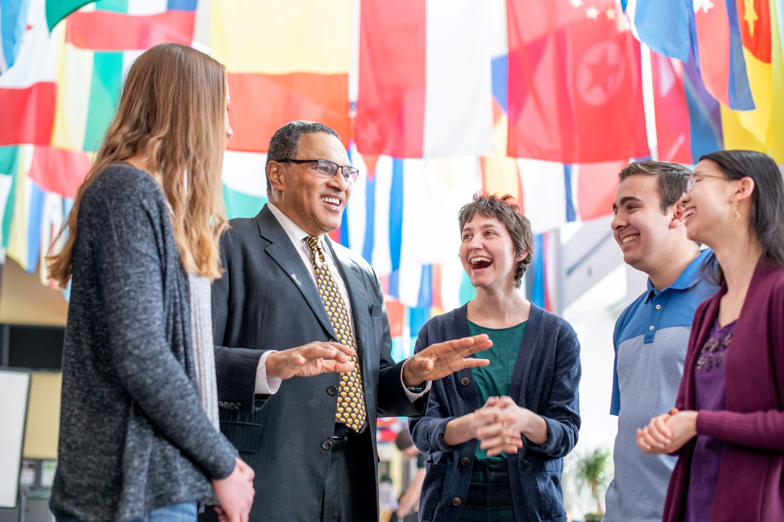 Older man in suit speaks with four smiling students, beneath international flags.