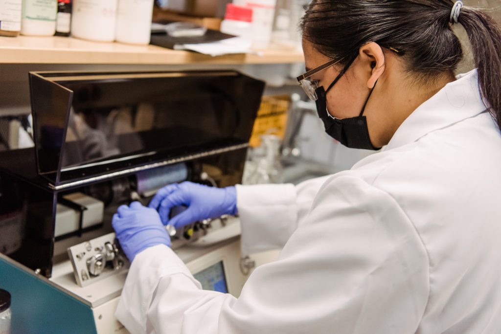 An Asian woman works in a science lab. She is wearing glasses, a black mask, a white lab coat, and blue gloves.