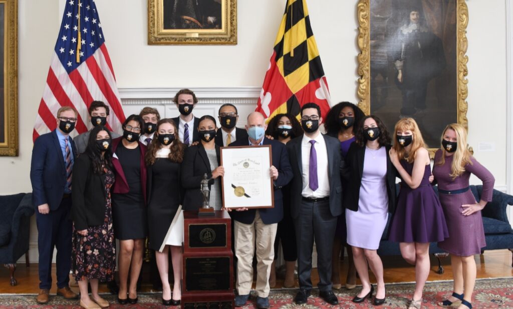 Over a dozen young adults and a few older adults in business attire stand in front of the US and Maryland flags, holding a framed document.