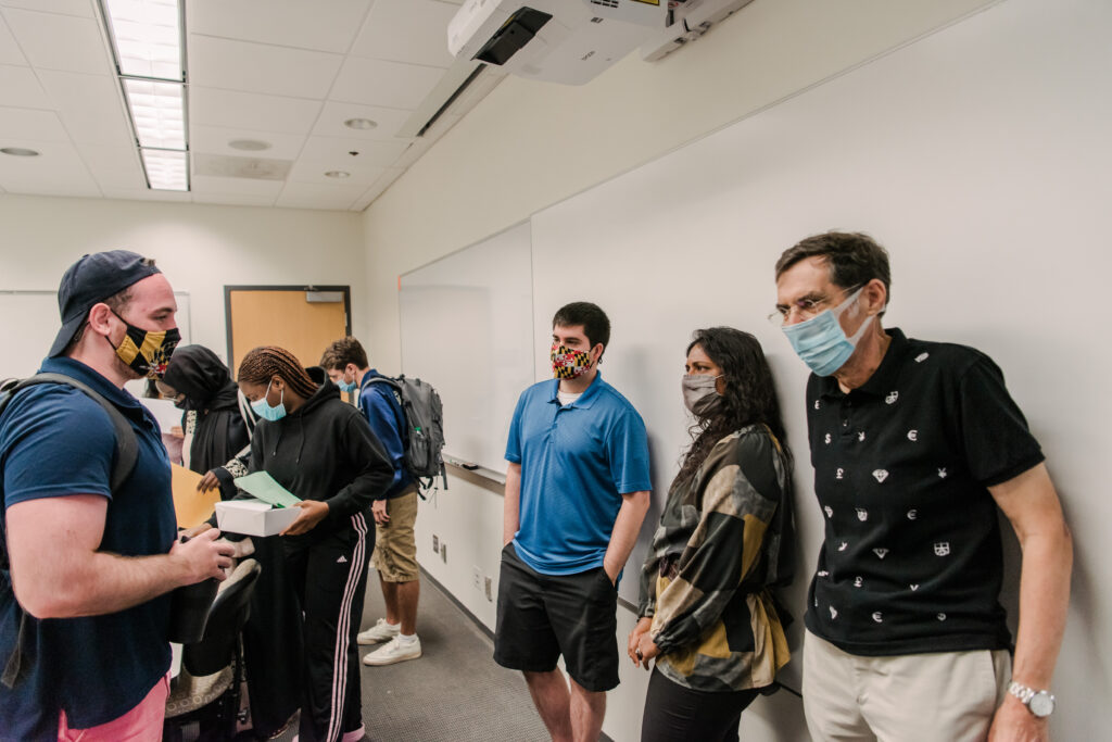 Two people wearing face masks talk to each other with numerous people and a white board in the background.