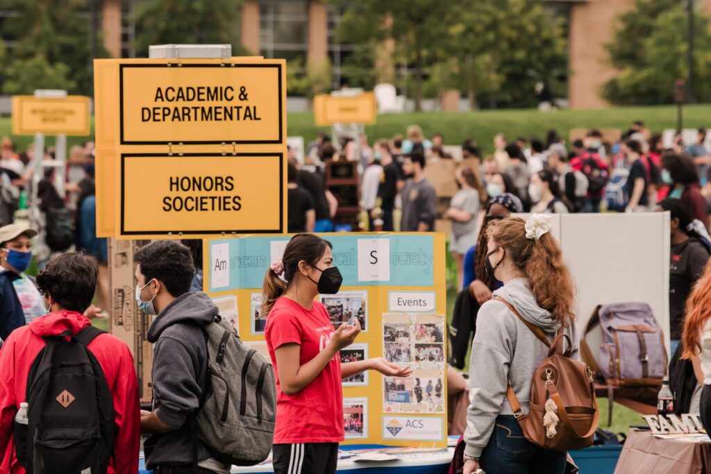 Two students wearing masks talk amidst a crowd of students outdoors. A sign reads Academic & Departmental and another reads Honors Societies.